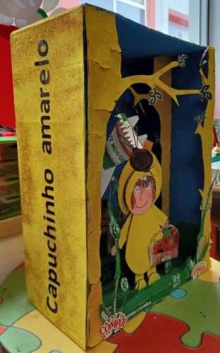 Capa - Elaborada com uma caixa de cartão pintada de amarelo com purpurinas por fora e de azul e verde por dentro. Com embalagens da TetraPak da marca Compal e algumas de leite da marca Agros construíram-se as personagens da história e alguns elementos par