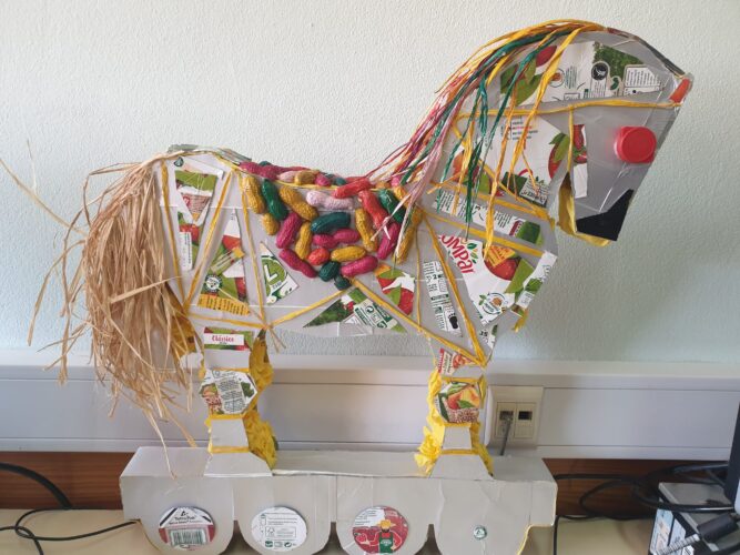 o símbolo é um cavalo de Tróia 3D, forrado com embalagens da Compal, restos de ráfia, cascas de amendoim e rolhas da mesma embalagem. São visíveis diversos símbolos.